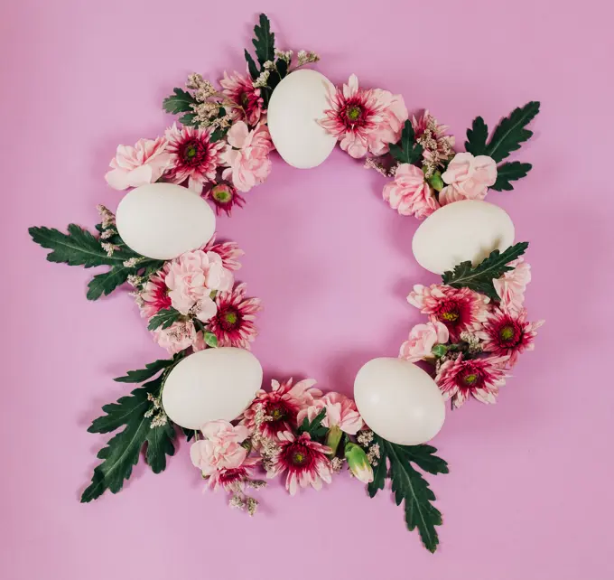 From above elegant Easter wreath made of flowers and chicken eggs and placed on pink background