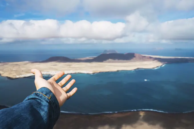 Hand of unrecognizable tourist showing small island in blue sea in Lanzarote, Spain.