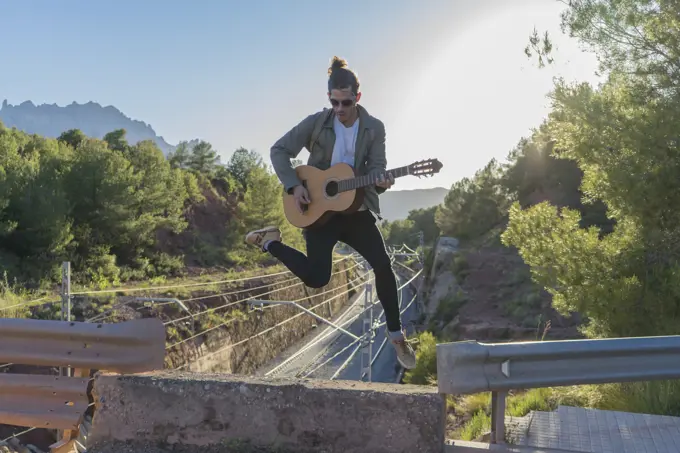 Young stylish man playing guitar in jump on background of landscape and railroad.