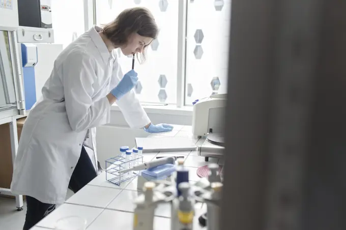 Side view of thoughtful woman standing with pen and working in modern laboratory.