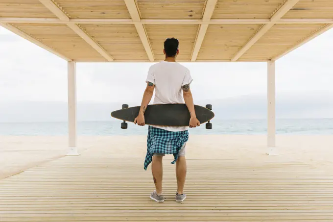 Tattooed man in summer apparel standing on shore with skateboard and looking at sea. Back view.