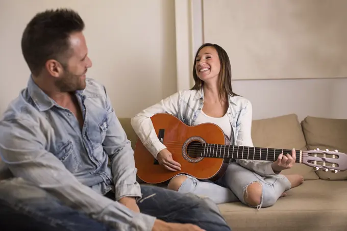 Cheerful young woman sitting on sofa and playing acoustic guitar for boyfriend at home.