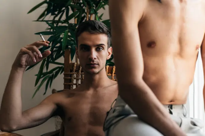 Stylish shirtless gay couple posing in front of the camera sitting in a chair smoking cigarette