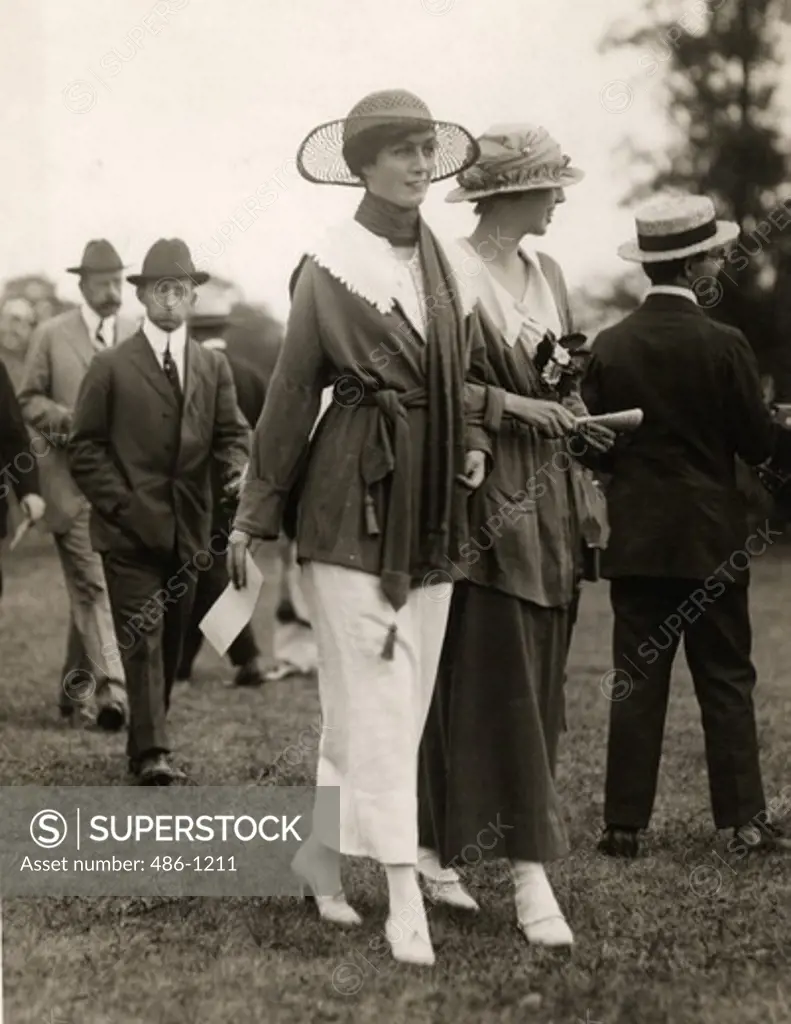 Two elegant women wearing hats walking on meadow, men in background