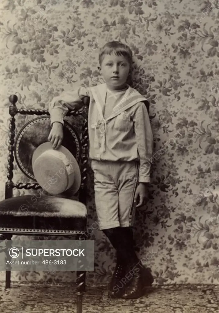 Portrait of boy posing with hat and leaning on chair, 1890