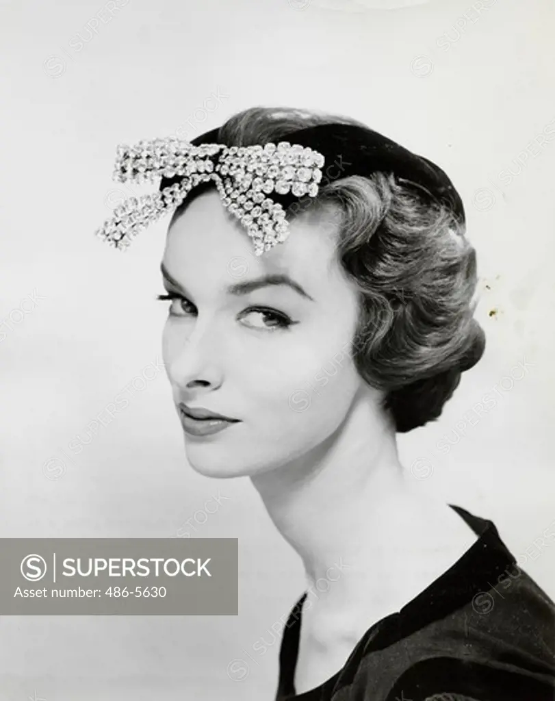Portrait of fashion model wearing evening headdress 1955, From Lilly Dache's Fall 1955 collection, ""glamour group"". This dramatic evening headdress is fashioned of black velvet in a squared cut-out design which reveals the coiffure. A bowknot of flashing brilliants is a headlight ornament