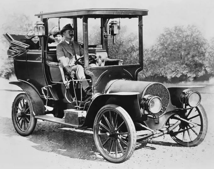Mature man driving a car with two mid adult women sitting behind him, 1908 Franklin