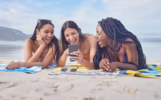 Girl friends, beach and phone of a teenager laughing at funny meme by the sea in Miami. Travel, vacation and sunshine with happy students enjoying spring break with mobile connection lying on sand