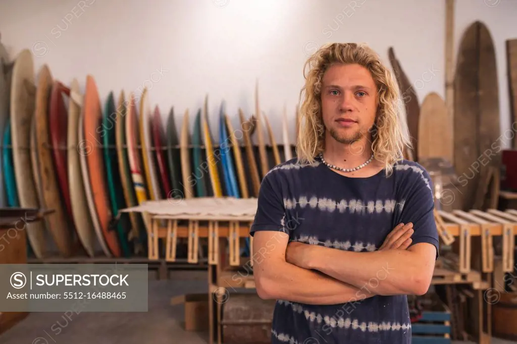 Portrait of a Caucasian male surfboard maker in his studio, with surfboards in a rack in the background, standing with his arms crossed and looking straight to camera. 
