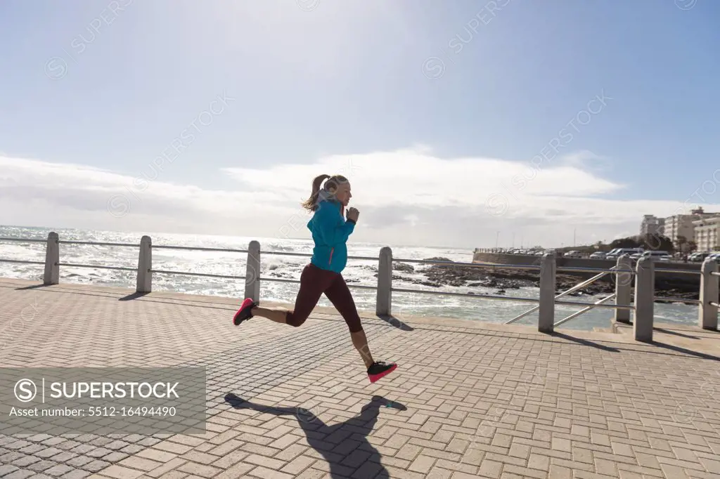 Side view of a sporty Caucasian woman with long dark hair exercising on a promenade by the seaside on a sunny day with blue sky, running.