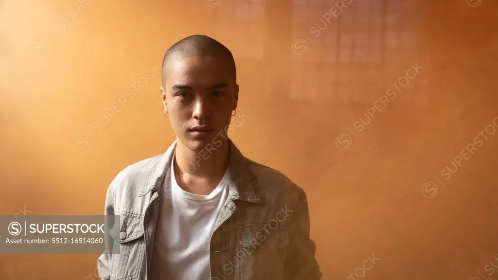 Front view of a young Hispanic-American man wearing a grey leather jacket over a white shirt looking intently at the camera inside an empty warehouse with orange smoke