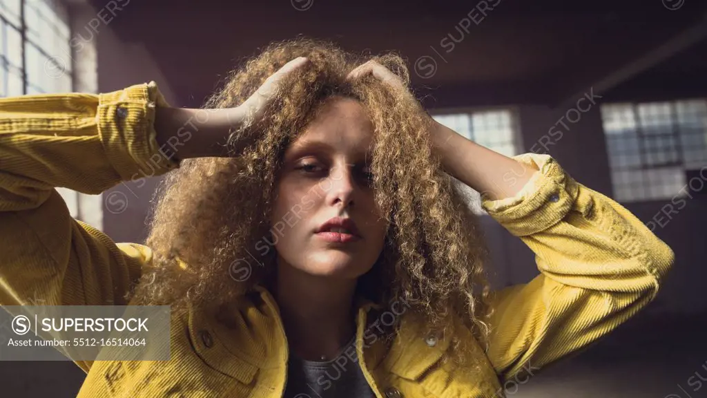 Front view of a young Caucasian woman with hands on curly hair wearing a yellow jacket over a grey shirt looking intently at the camera inside an empty warehouse