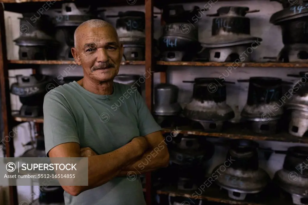 Portrait of a smiling senior mixed race man with arms crossed, standing in front of shelves of traditional equipment in the workshop at a hat factory