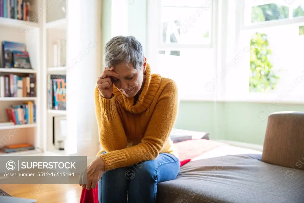 Front view close up of a mature Caucasian woman with short grey hair sitting at home in her living room looking down with her head leaning on her hand, a sunlit window in the background
