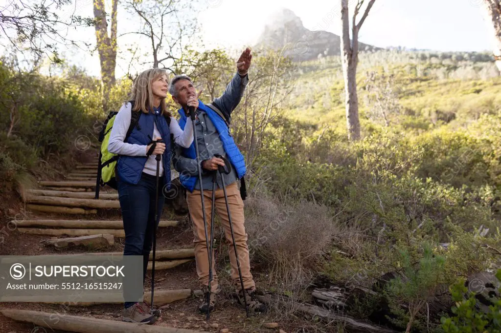 Front view of a mature Caucasian woman and man wearing backpacks and using Nordic walking sticks, stopping on a downhill trail to admire the view and pointing to the distance during a hike in the countryside