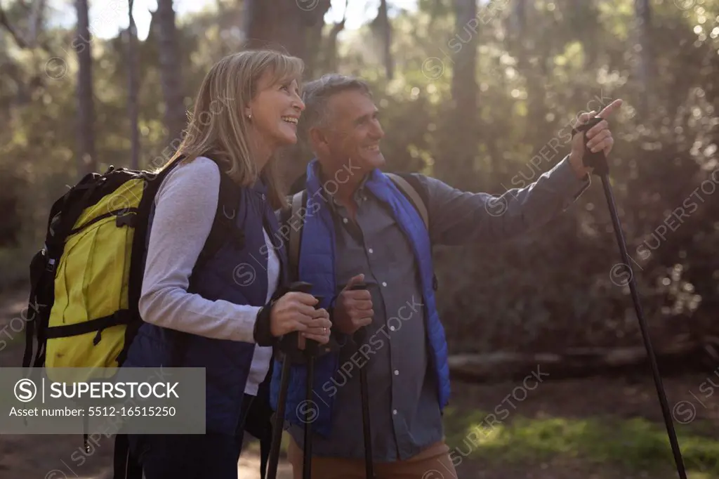 Side view close up of a mature Caucasian woman and man wearing backpacks and using Nordic walking sticks, stopping on a trail to admire the view and pointing to the distance during a hike in the countryside