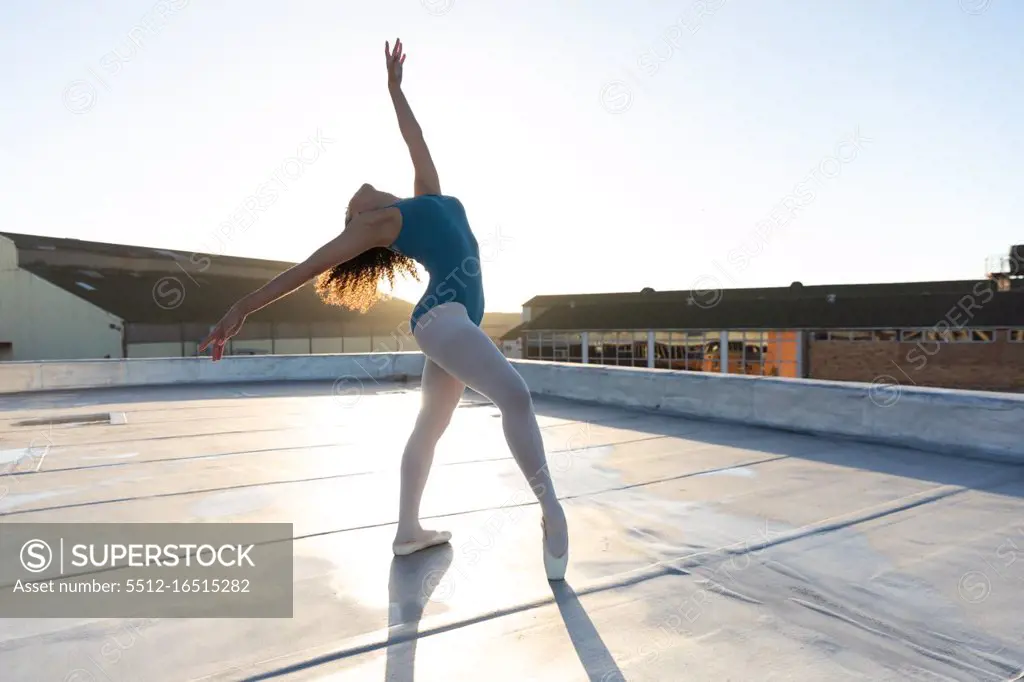 Side view of a young mixed race female ballet dancer leaning back in a ballet pose with arms oustretched, on the rooftop of an urban building, backlit by sunlight
