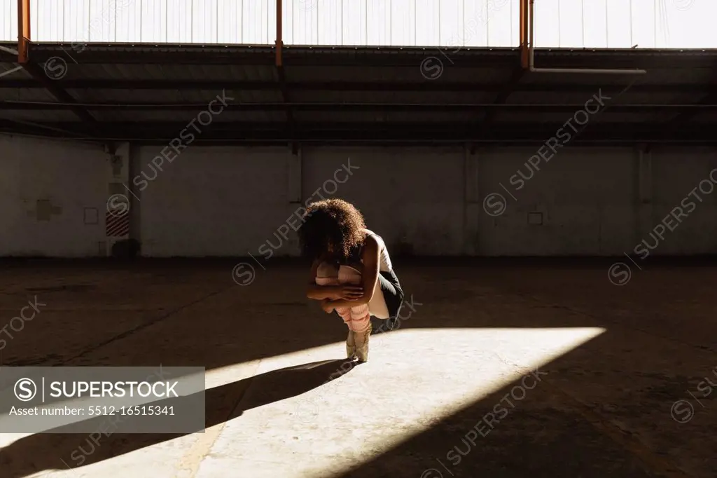 Front view of a young mixed race female ballet dancer wearing pointe shoes squatting down holding her knees balanced on her toes in shaft of sunlight while dancing in an empty room at an abandoned warehouse