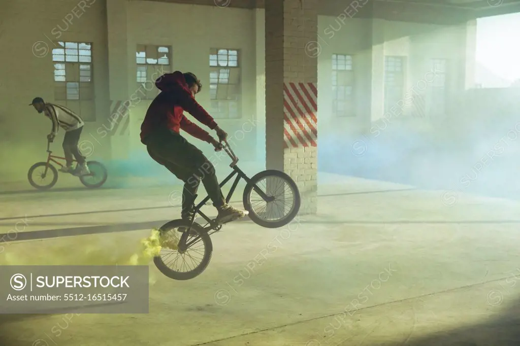 Side view of two young Caucasian men riding and doing tricks on BMX bikes with yellow and blue smoke grenades attached to them, in an abandoned warehouse