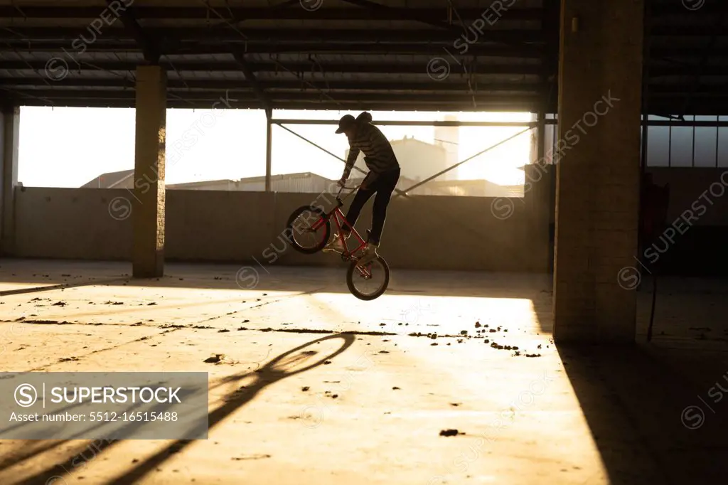 Side view of a young Caucasian man jumping on a BMX bike while practicing tricks in an abandoned warehouse, backlit by sunlight