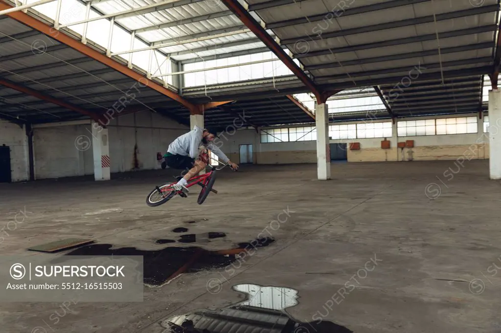 Side view of a young Caucasian man riding a BMX bike, jumping off the ground and turning the handlebars, while practicing tricks in an abandoned warehouse