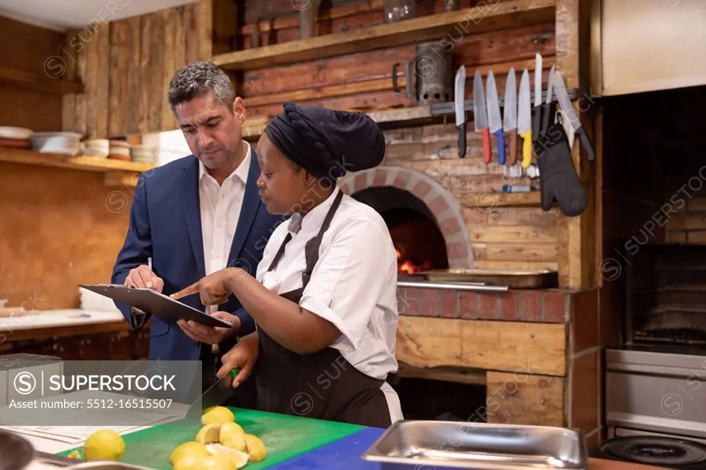 Front view close up of a middle aged Caucasian restaurant manager holding a clipboard and talking with a young African American female chef in a restaurant kitchen