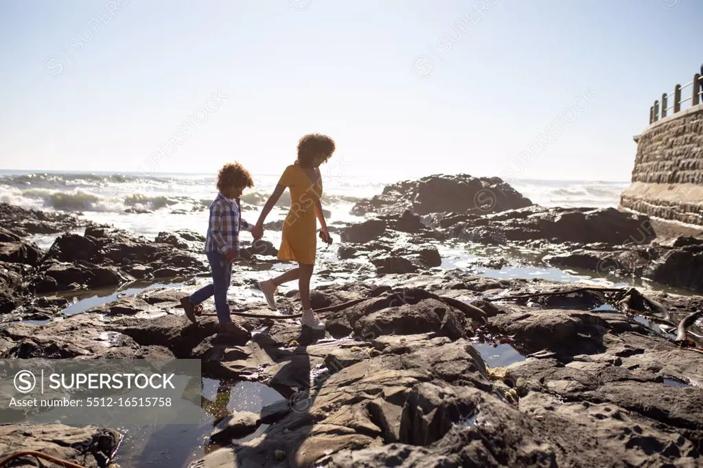 Side view of a young mixed race woman and her pre-teen son enjoying time together by the sea, holding hands and walking on a sunny day