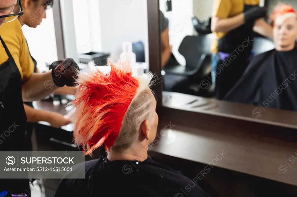 Rear view of a middle aged Caucasian male hairdresser and a young Caucasian woman having her hair coloured bright red in a hair salon, reflected in a mirror