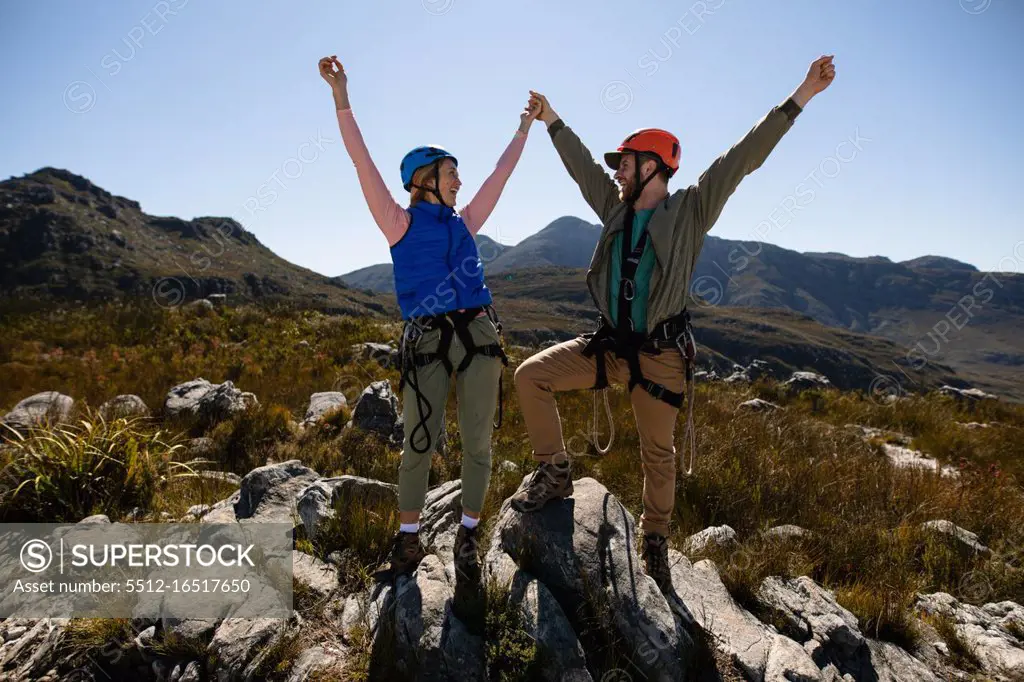 Front view of Caucasian couple enjoying time in nature, wearing zip lining equipment, holding hands with arms in the air, smiling at each other on a sunny day in mountains