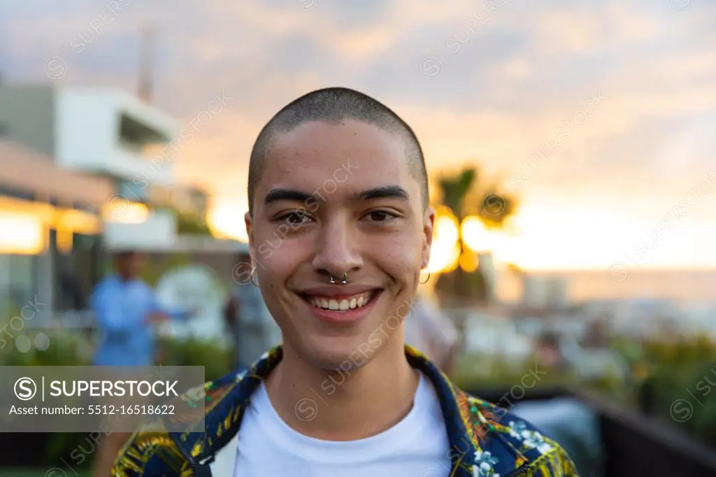 Portrait of a mixed race man hanging out on a roof terrace with a sunset sky, looking at camera and smiling