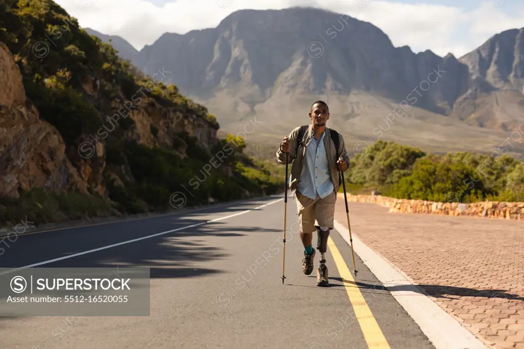 A fit, disabled mixed race male athlete with prosthetic leg, enjoying his time on a trip to the mountains, hiking with sticks, walking on the road in the mountains. Active lifestyle with disability.