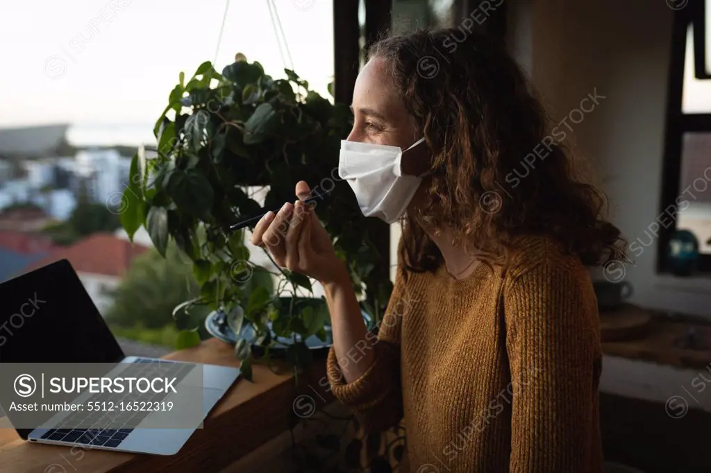 Caucasian woman spending time at home self isolating and social distancing in quarantine lockdown during coronavirus covid 19 epidemic, wearing a face mask against covid19 coronavirus, standing by a window, talking on her smartphone and using a laptop computer.