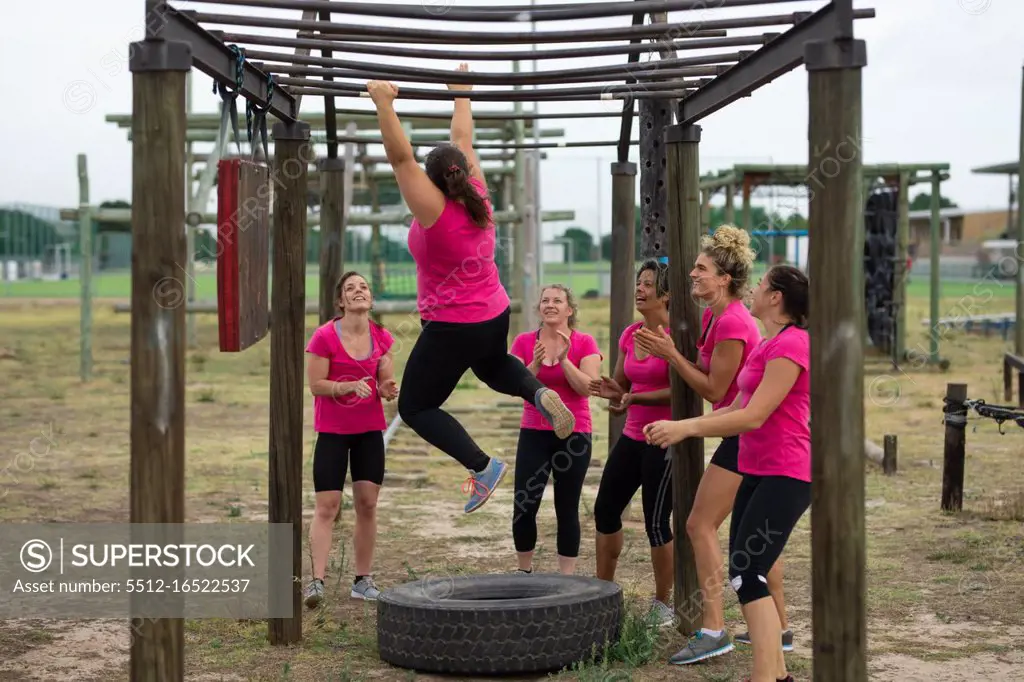 Multi-ethnic group of women all wearing pink t shirts at a boot camp training session, exercising, hanging from monkey bars. Outdoor group exercise, fun healthy challenge.