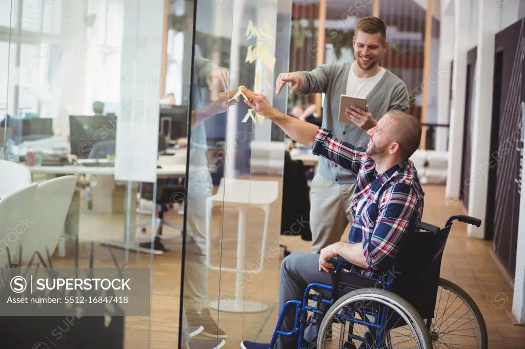 Businessman assisting handicap colleague while sticking adhesive notes on glass window in creative office
