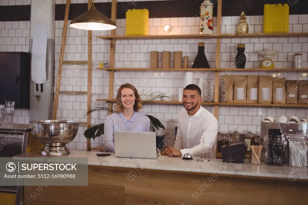 Portrait of smiling waitress and male owner using laptop at counter in coffee shop