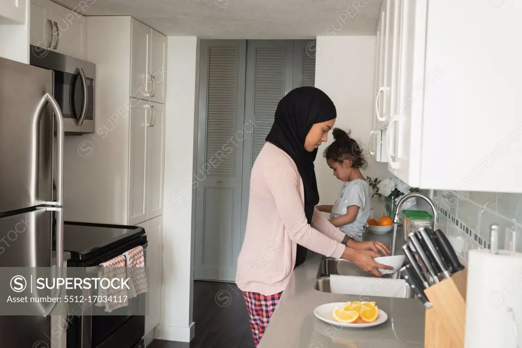 Side view of mixed race mother wearing hijab working in kitchen room while daughter sitting behind her at home