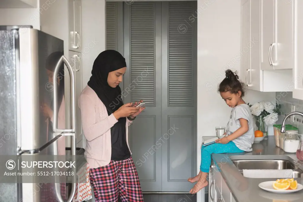 Side view of mixed race mother wearing hijab using mobile phone while standing in kitchen room at home