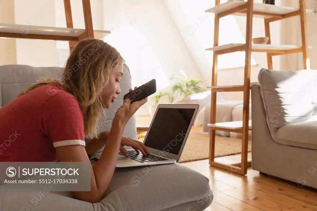Side view of caucasian woman using laptop while talking on mobile phone in living room at home 