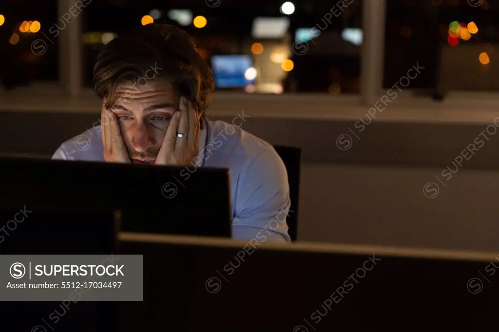 Front view of tired young Caucasian male executive with hands on face working at desk in a modern office. He is working late
