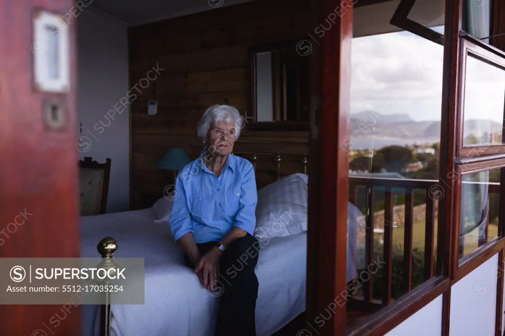  Front view of a thoughtful active senior woman sitting on the bed and looking through the window in bedroom at home