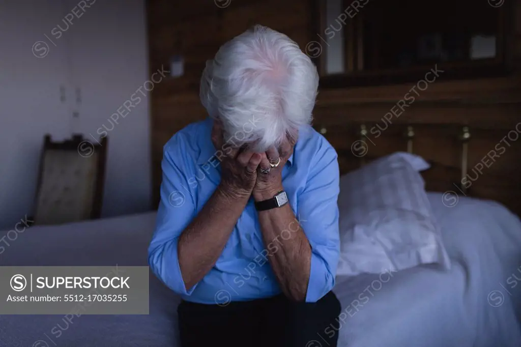 Front view of a sad active senior woman covering her face with her hands while sitting on the bed in bedroom at home