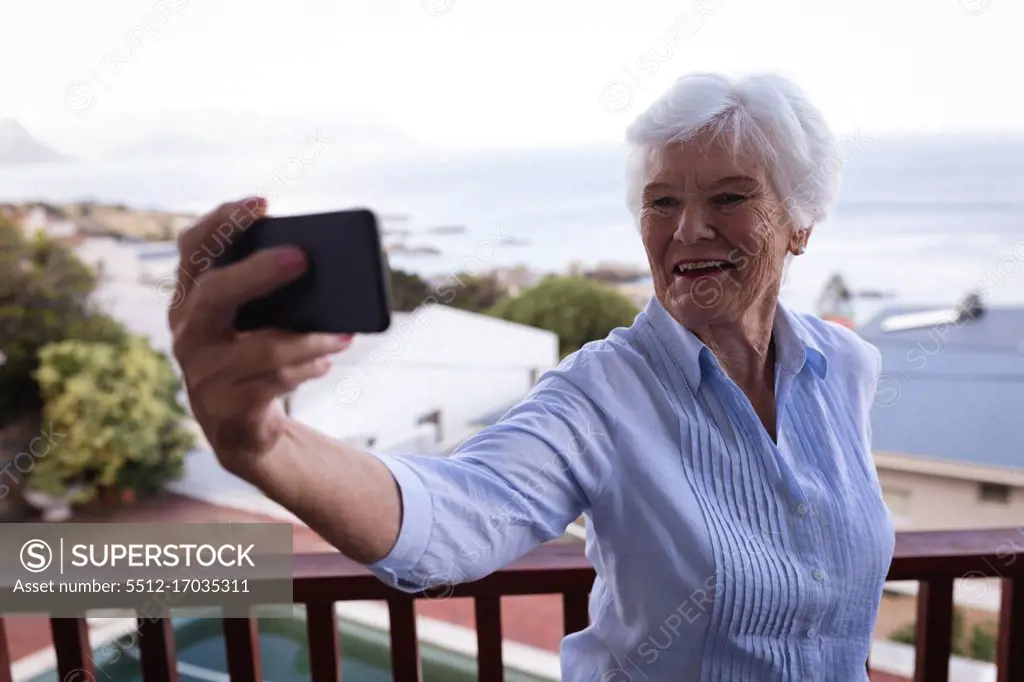 Front view of a happy active senior woman taking a selfie on the balcony against a sea landscape with her mobile phone at home