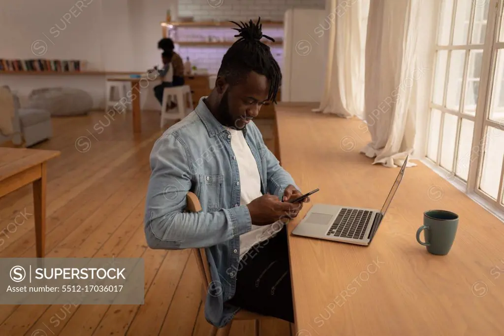 Side view of man using mobile phone with laptop on table at home