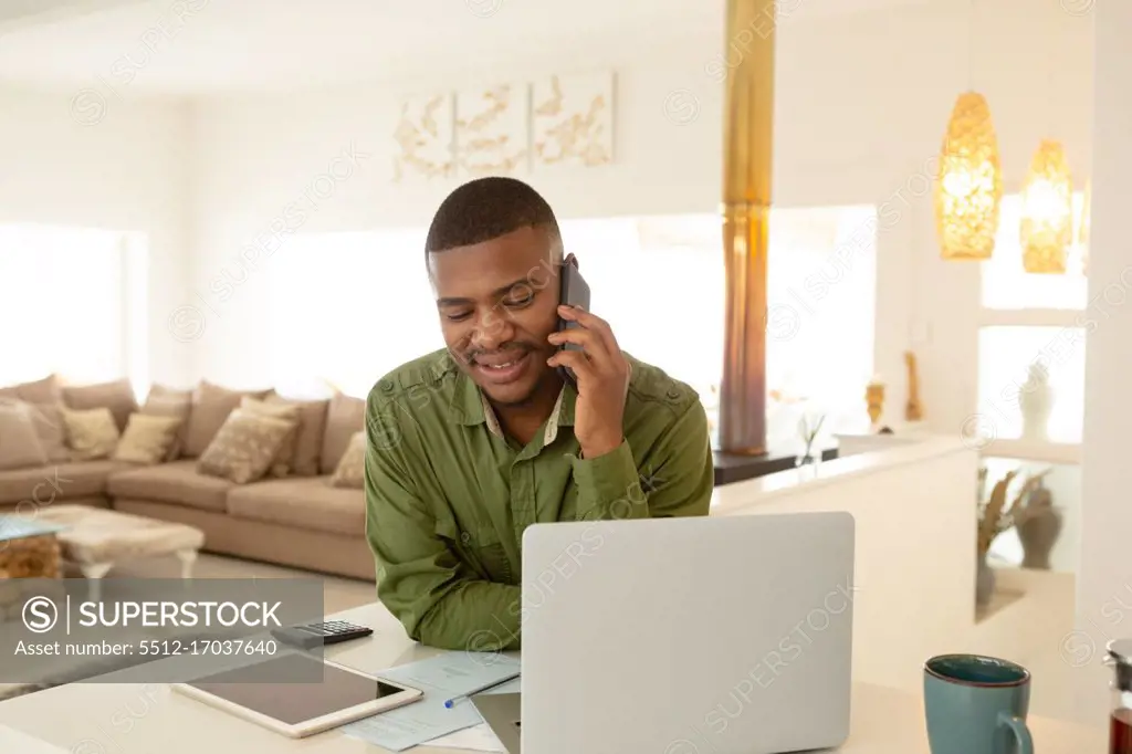 Front view of happy African-american man talking on mobile phone at table in a comfortable home. Authentic home lifestyle setting with young African American male
