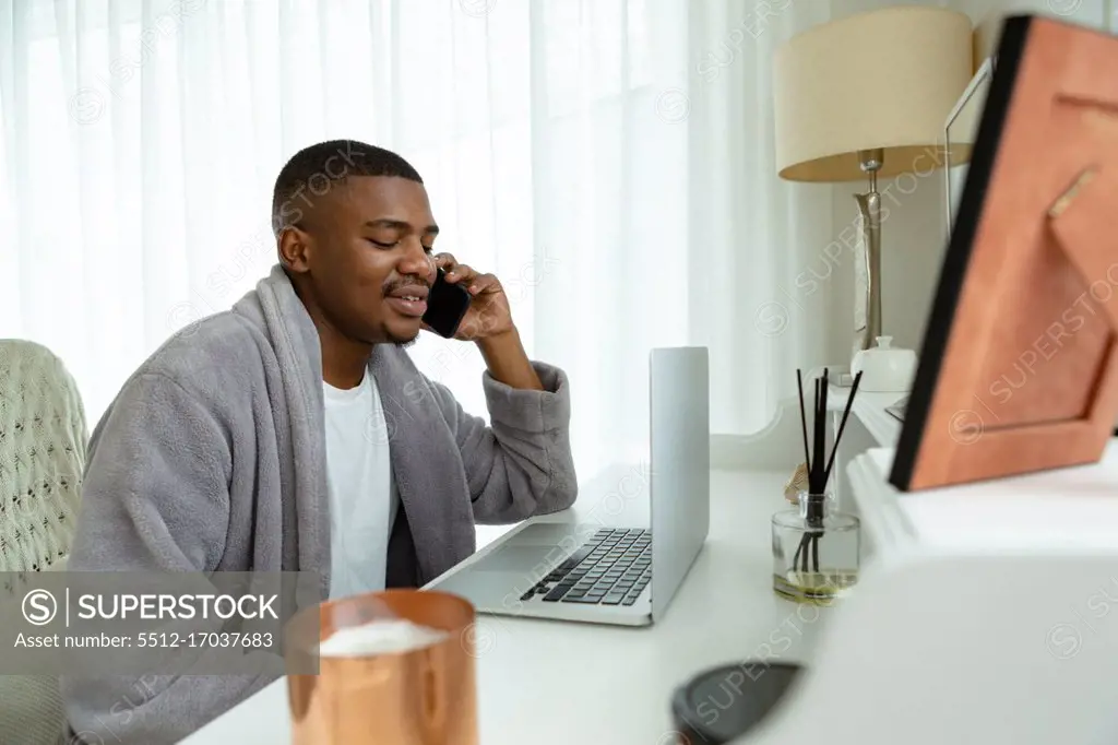 Side view of African-american man talking on mobile phone while working on laptop at desk in bedroom at comfortable home. Authentic home lifestyle setting with young African American male