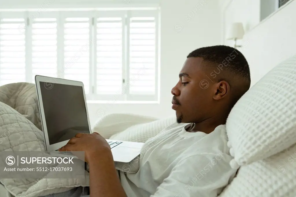 Side view of African-american man using laptop while lying on bed in bedroom at comfortable home. Authentic home lifestyle setting with young African American male