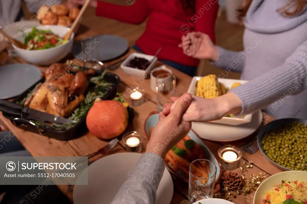 High angle view of a young mixed race man and two young adult Caucasian women sitting at a table holding hands saying grace before eating Thanksgiving dinner at home together with friends
