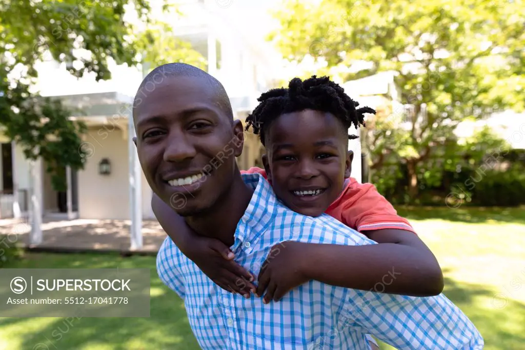 Self isolation in quarantine lock down. portrait of an african american man outside outside his house in the garden on a sunny day, piggybacking his young son, both smiling to camera