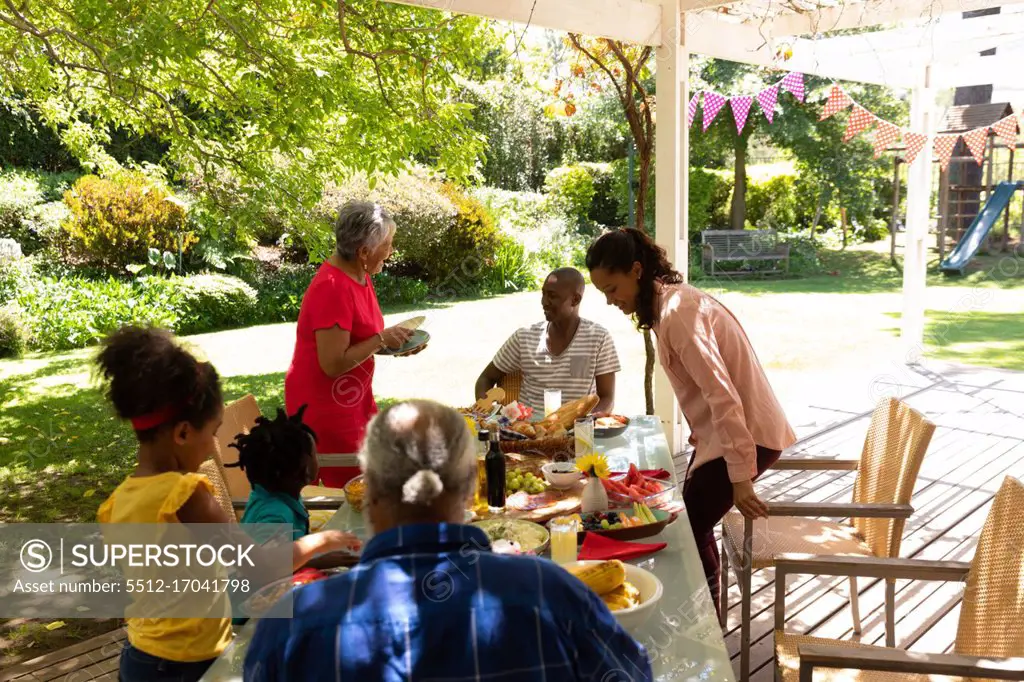 Side view of a multi-ethnic, multi-generation family serving food and sitting down at a table for a meal together outside on a patio in the sun