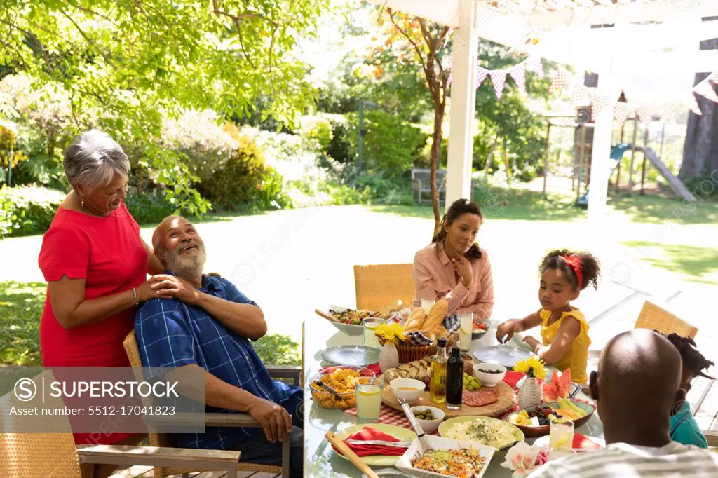 Side view of a multi-ethnic, multi-generation family sitting at a table for a meal together outside on a patio in the sun, the grandparents embracing, the grandmother standing behind the grandfather
